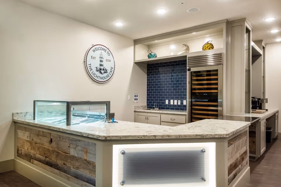 Modern café-style service counter with marble countertops, a refrigerated display case, wine fridge, and a wall sign reading 'Welcome to The Lighthouse Cafe'.