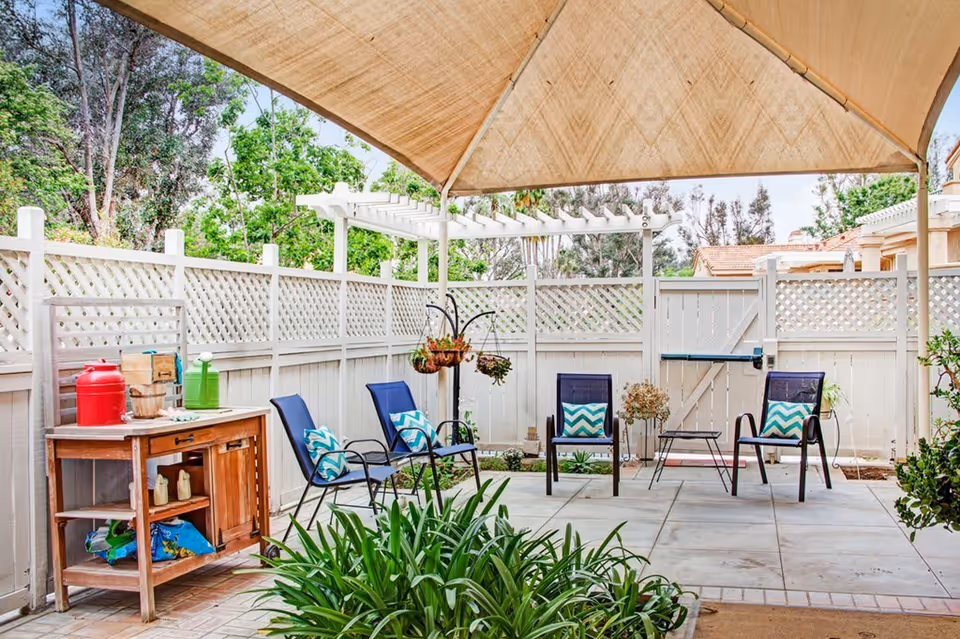 Outdoor patio area with a beige canopy overhead, four blue chairs with patterned cushions arranged around a small table, a wooden side table with various items on it, and green plants in the foreground and background. The area is enclosed by a white lattice fence with trees visible beyond.