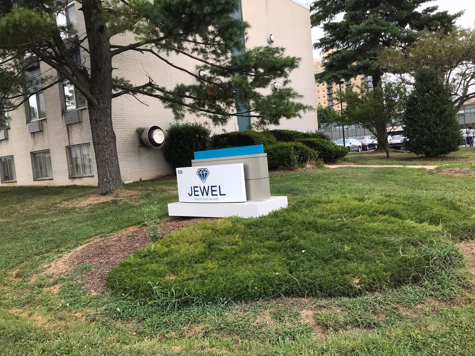 Outdoor view of a building with a sign in front that reads 'JEWEL Health and Rehab' surrounded by grass, bushes, and trees.