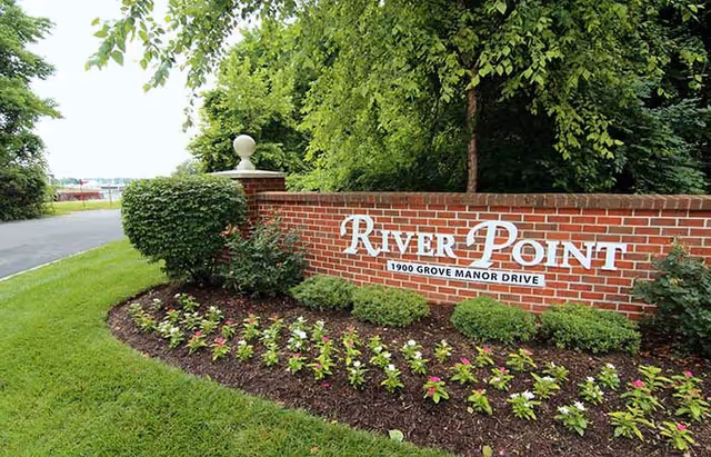 A brick sign for River Point with the address 1900 Grove Manor Drive, surrounded by neatly trimmed bushes and a flower bed, with trees and a road visible in the background.
