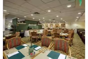 Spacious dining room with multiple tables and chairs arranged neatly. The tables are set with placemats, napkins, and small decorative items. The room has a carpeted floor, a drop ceiling with recessed lighting, and a green accent wall with shelves and decorative items.