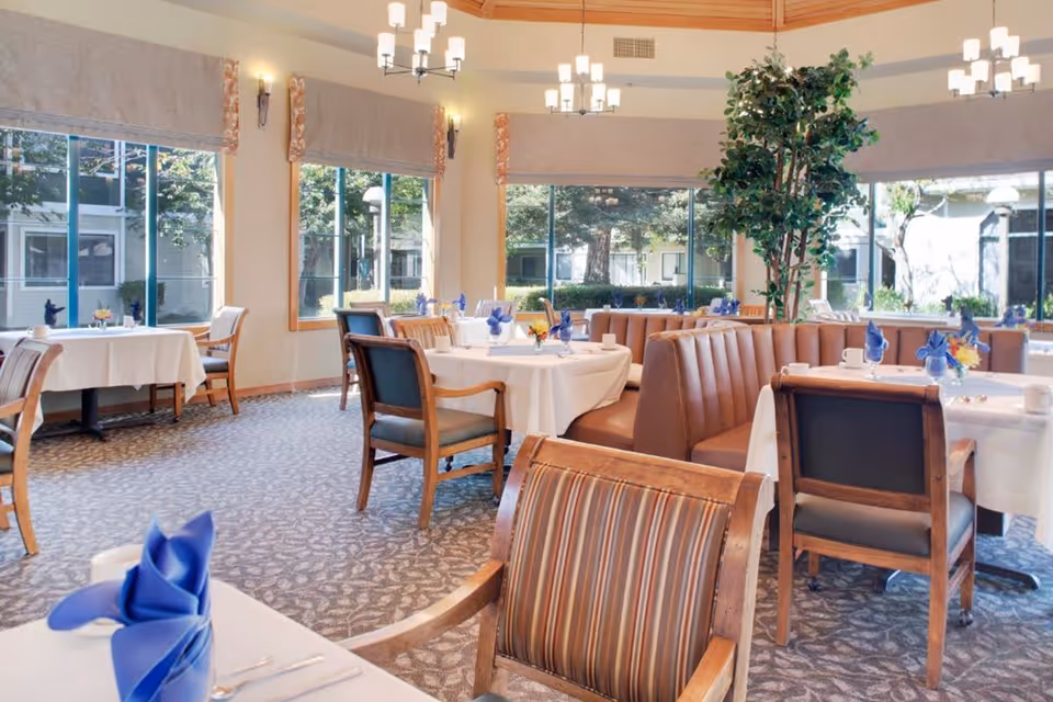 Bright dining room with large windows showing outdoor greenery. Tables are set with white tablecloths, blue folded napkins, and small flower arrangements. The room features a mix of wooden chairs and brown cushioned booth seating under warm ceiling lights.