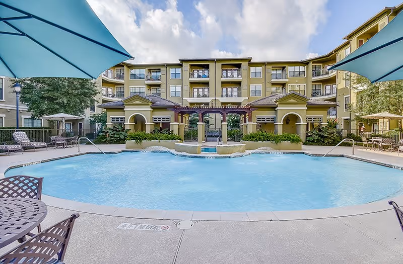 Outdoor swimming pool area with lounge chairs, tables with umbrellas, and a multi-story residential building in the background under a partly cloudy sky.