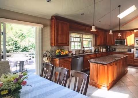 Bright kitchen with wooden cabinets and a central island, dining table and chairs in the foreground and a sliding glass door to a patio.