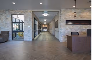 Interior view of a senior living facility hallway with stone accent walls, large windows on the left, a wooden reception desk on the right, and modern ceiling lights.