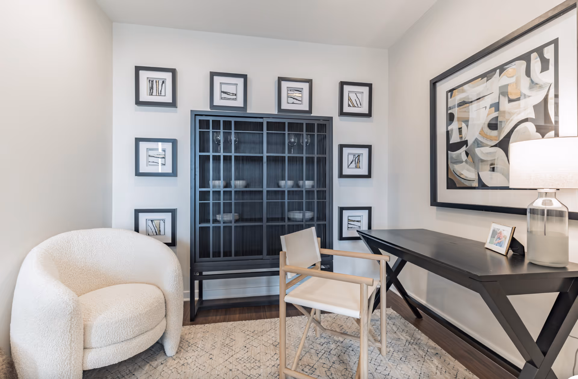 A cozy interior room featuring a white upholstered armchair, a black wooden cabinet with glass doors displaying dishes, a black desk with a lamp and a framed photo, and abstract artwork on the wall. The walls are decorated with a series of small framed pictures arranged around the cabinet.