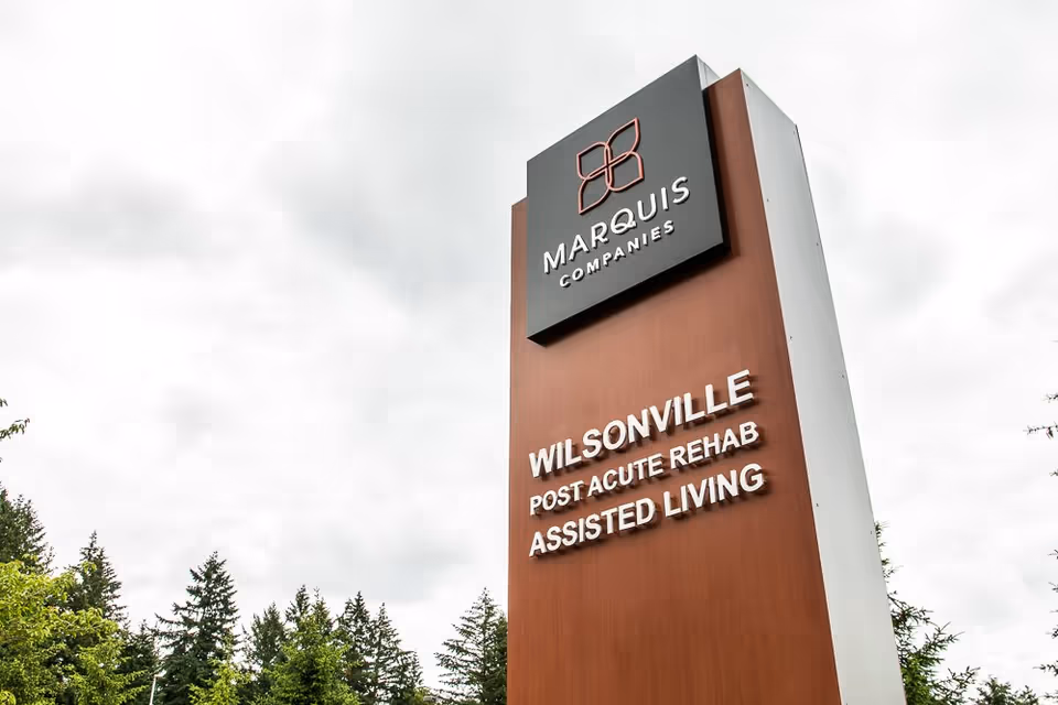 Tall outdoor sign for Marquis Companies in Wilsonville, indicating Post Acute Rehab and Assisted Living services, with trees and a cloudy sky in the background.