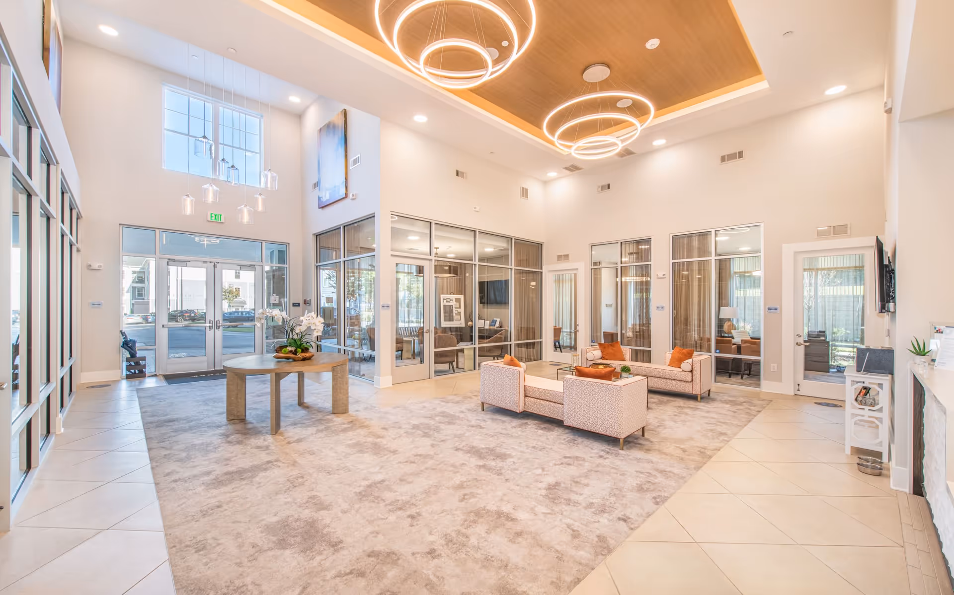 Bright and spacious senior living facility lobby with high ceilings, modern circular pendant lights, a central round table with a floral arrangement, and two seating areas with patterned sofas and orange cushions. Glass walls and doors lead to adjacent rooms, and large windows allow natural light to fill the space.