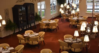 Interior view of a dining room with multiple round tables set for meals, surrounded by beige upholstered chairs. The room features large windows letting in natural light, a dark wooden cabinet against the wall, decorative wall art, and several chandeliers with lampshades providing warm lighting. The carpet is a deep red color.