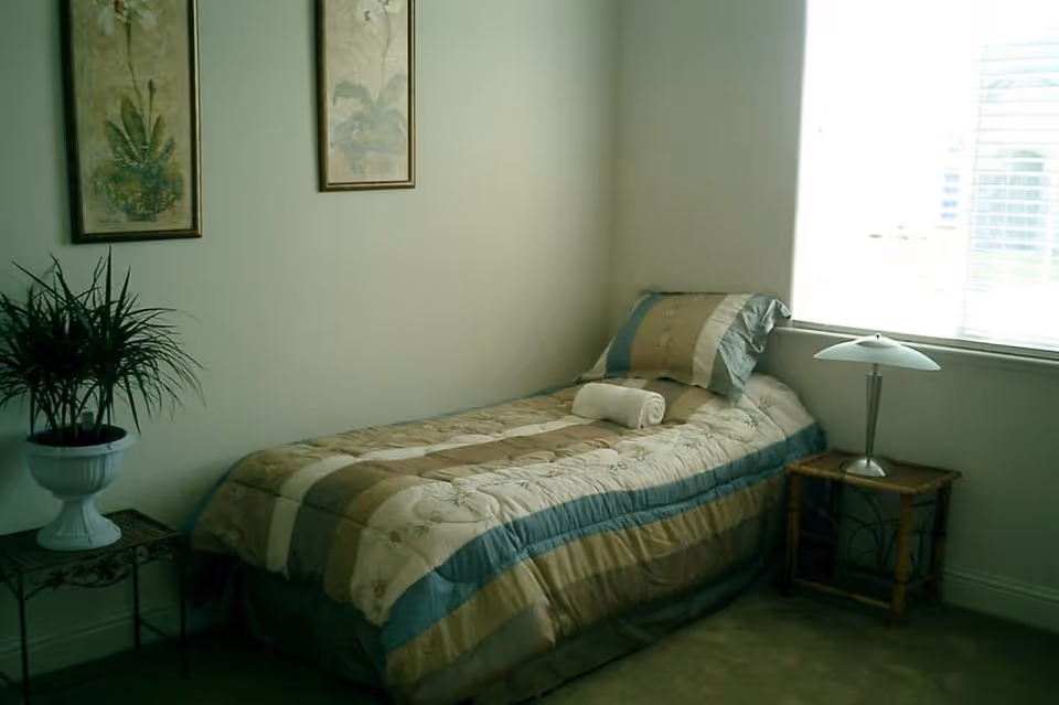 A small bedroom with a single bed covered in a beige, blue, and green striped quilt. A rolled white towel is placed on the bed. Next to the bed is a small wooden nightstand with a modern lamp. On the opposite side, there is a small table with a potted plant. Two framed botanical prints hang on the wall above the bed. A window with blinds allows natural light into the room.