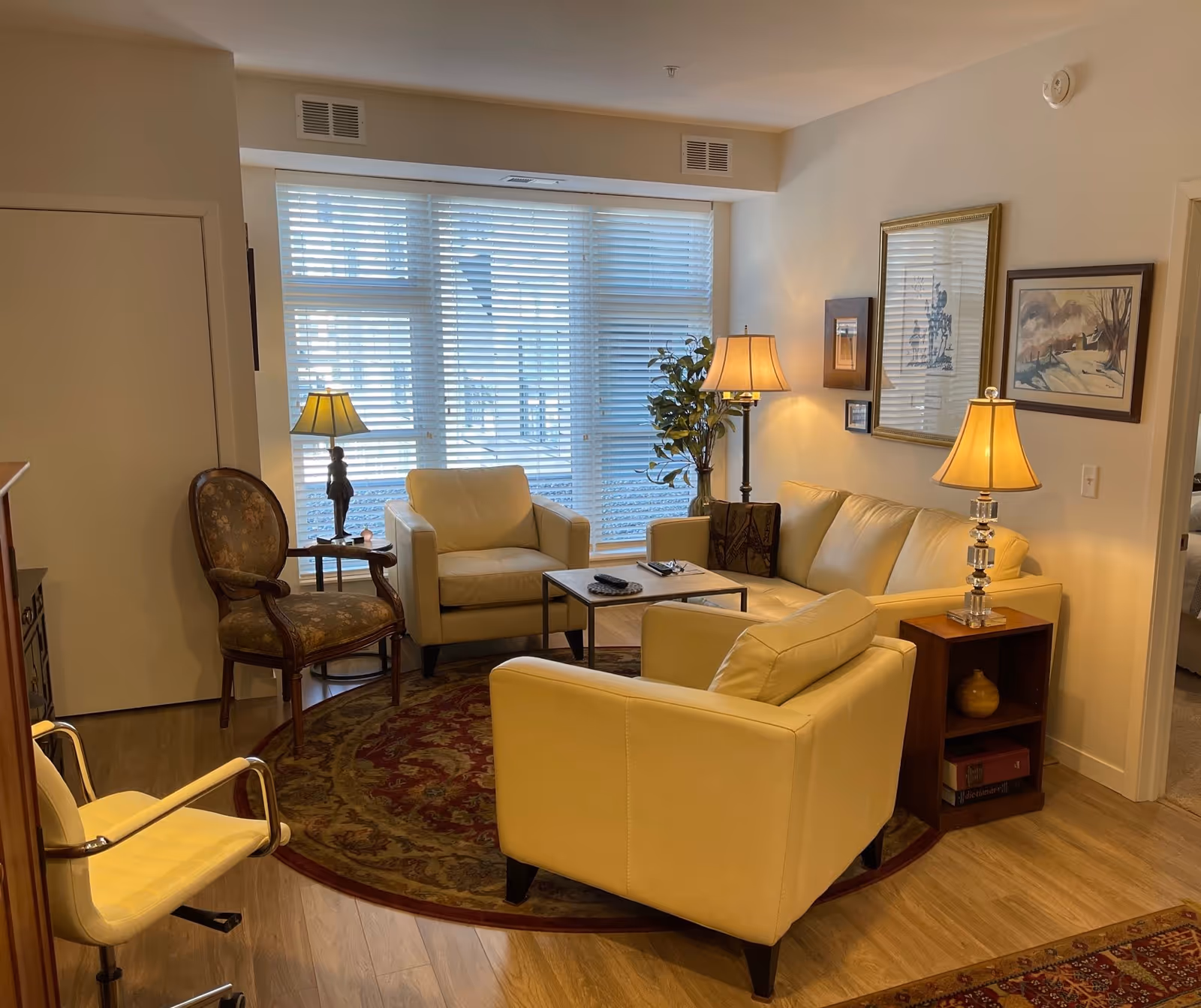A cozy living room with cream-colored leather sofa and armchairs arranged around a small wooden coffee table on a round patterned rug. The room has large windows with white blinds, two table lamps, a floor lamp, framed artwork on the walls, and a wooden side table with books and a decorative item.