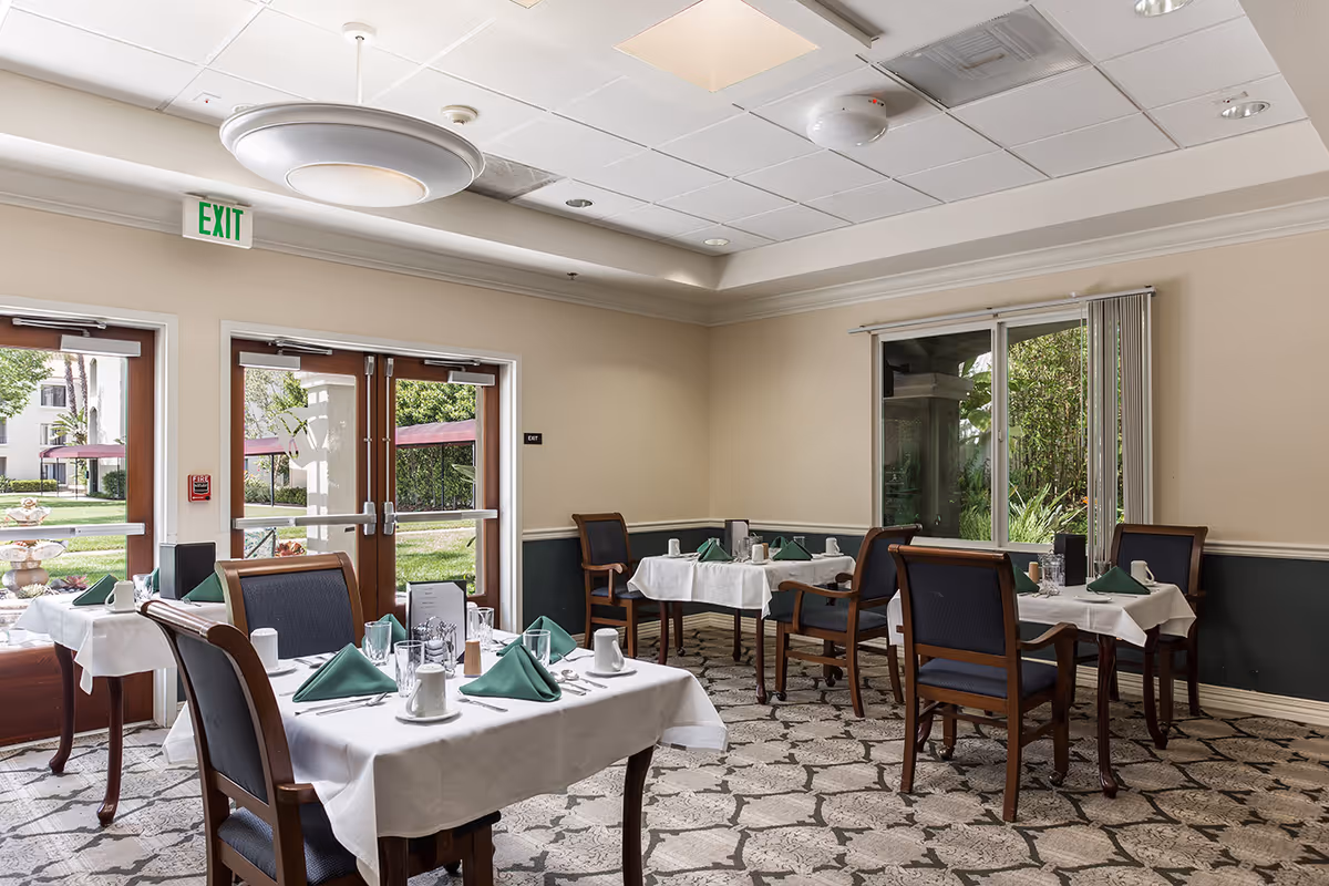Dining room with several tables covered with white tablecloths, each set with green folded napkins, cups, glasses, and silverware. The room has large windows and glass doors that open to a garden area outside. The ceiling has recessed lighting and a large circular light fixture.