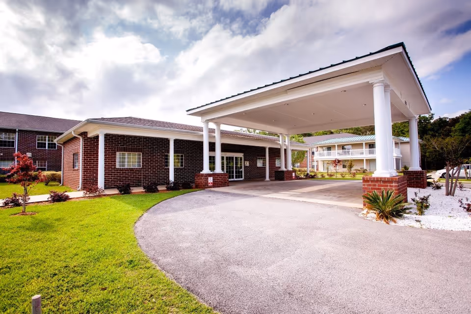 Entrance of a brick senior living facility featuring a large covered porte-cochere, driveway, and landscaped lawn.