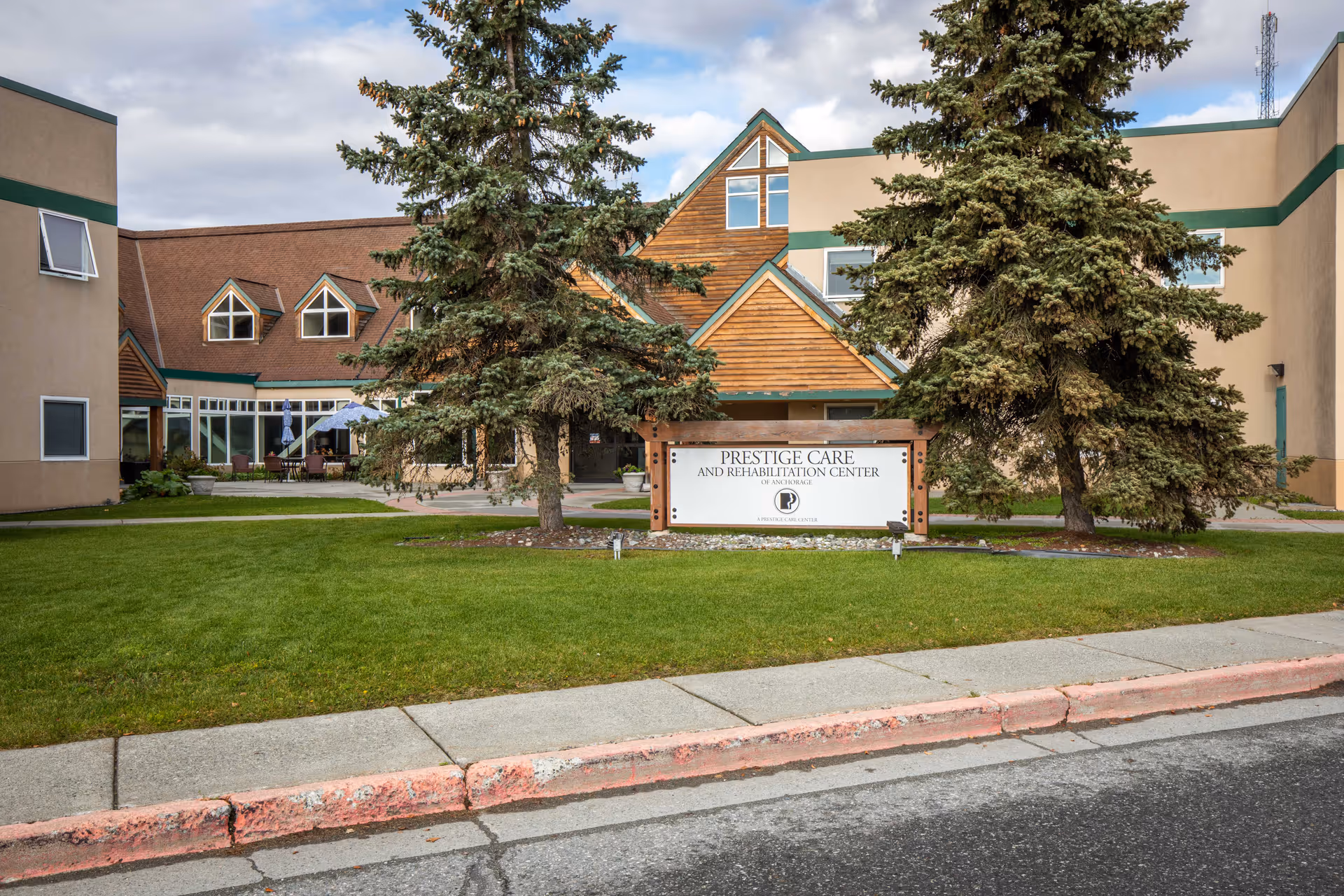 Front exterior of a rehabilitation center with a lawn, two large evergreen trees, and a sign in front of the building.