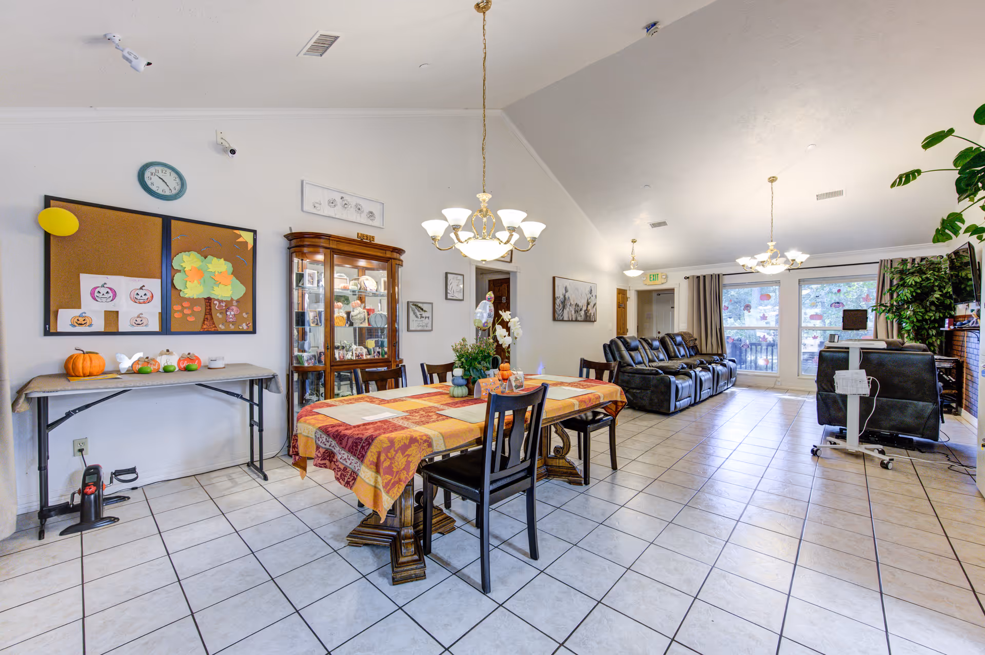 A spacious senior living common area with a dining table covered by a colorful tablecloth and surrounded by chairs. On the left wall, there is a bulletin board with autumn-themed decorations and a small table with pumpkins. A glass display cabinet with various items is next to the bulletin board. The room has tiled floors, multiple ceiling lights, and a seating area with black recliners near large windows with curtains. There is a TV mounted on the right wall and some plants adding greenery to the space.