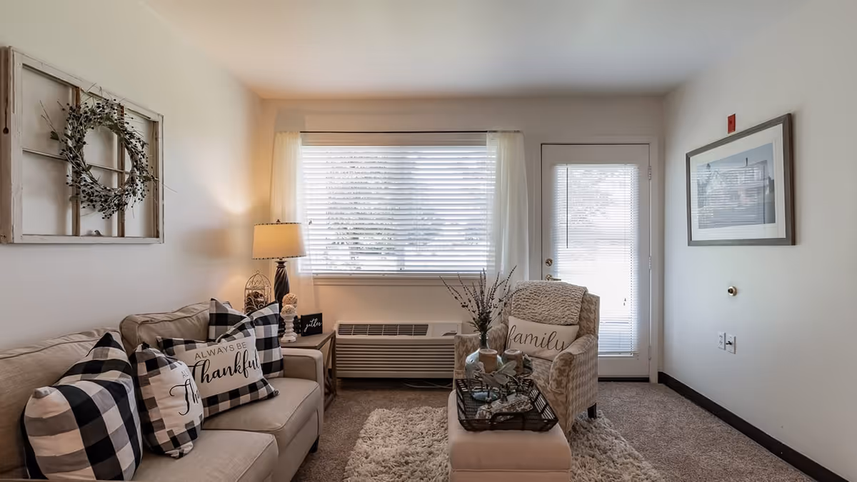 A cozy living room with a beige sofa adorned with black and white checkered pillows, one reading 'Always be thankful'. Next to the sofa is a side table with a lamp and decorative items. Across from the sofa is a beige armchair with a pillow that says 'family', positioned next to a glass door with blinds. A window with white curtains and blinds is centered on the back wall above a heating/cooling unit. The room has a soft carpet and neutral-colored walls decorated with a framed picture and a rustic window frame with a wreath.