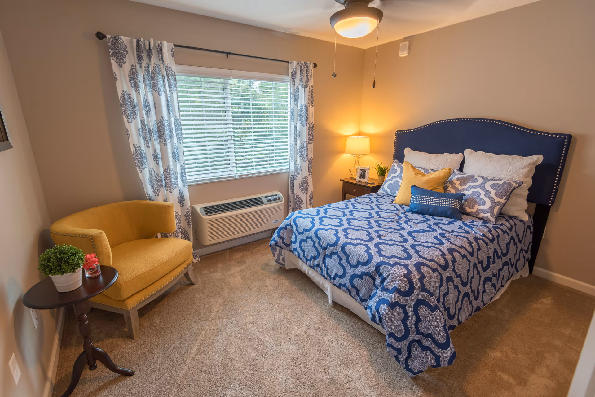 A cozy bedroom with a blue patterned bedspread on a bed with a blue upholstered headboard. There are multiple pillows on the bed including white, yellow, and blue ones. A yellow armchair is positioned next to a small round wooden table with two potted plants. The room has beige walls, a window with white blinds and patterned curtains, a ceiling fan with a light, and a nightstand with a lamp and picture frame.