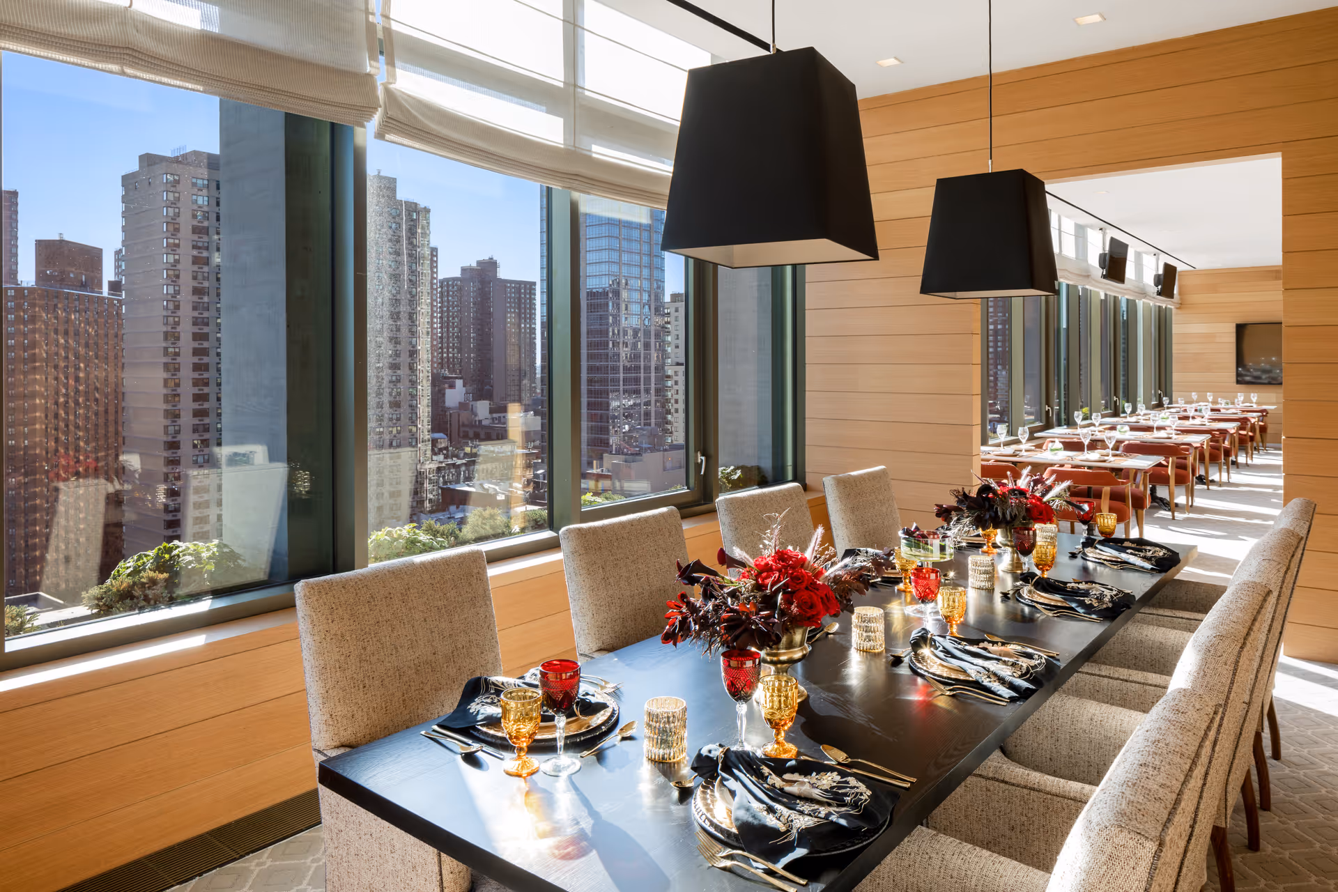 A bright dining room with a long black table set for a meal, featuring beige upholstered chairs, red and amber glassware, black and gold napkins, and floral centerpieces. Large windows offer a view of city buildings outside, and modern black pendant lights hang above the table. Another dining area with similar setup is visible in the background.
