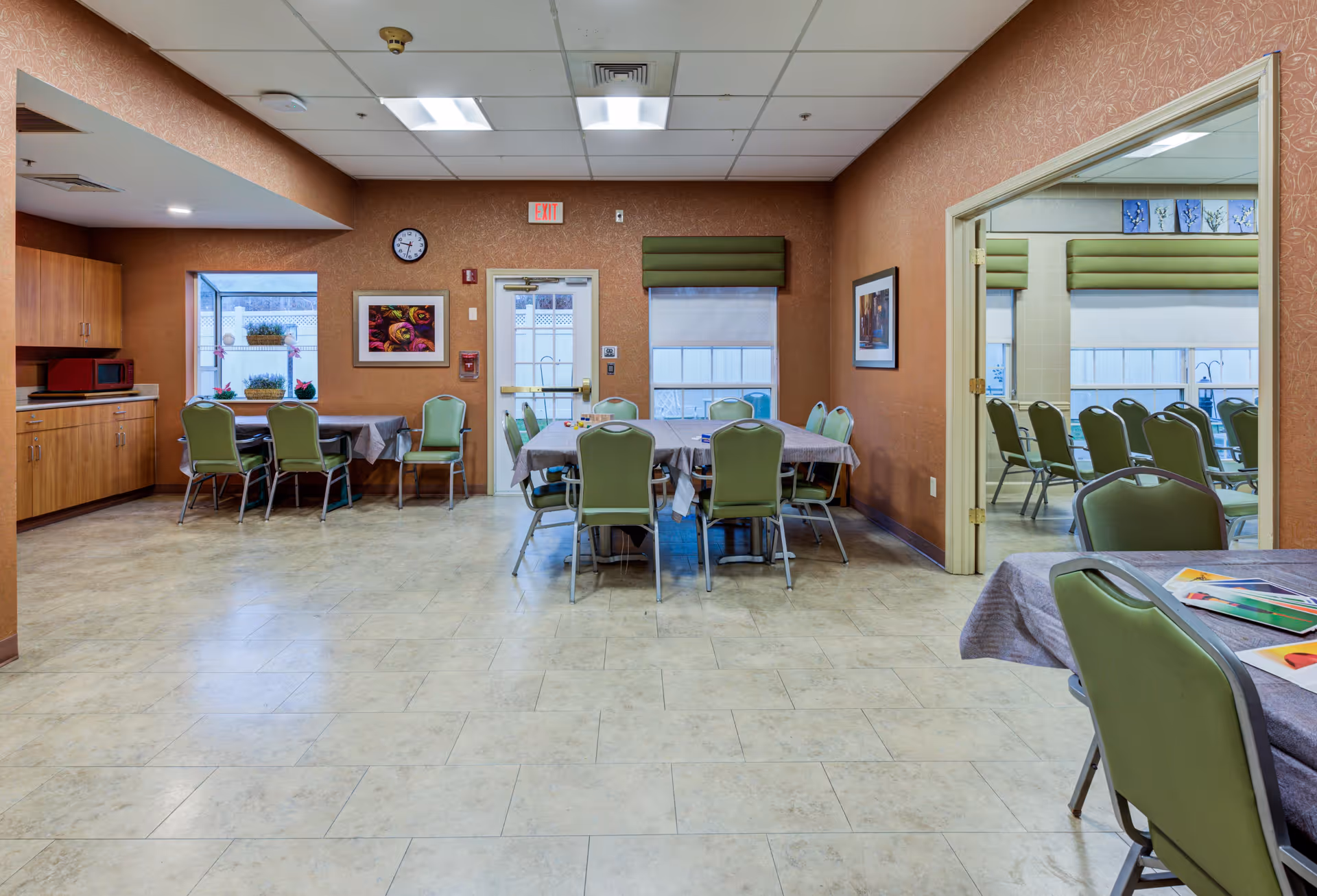 Interior view of a dining room in a senior living facility with multiple tables covered with gray tablecloths and green chairs around them. There is a small kitchenette area with wooden cabinets and a microwave on the left side. The walls are decorated with framed pictures and a clock above the exit door. The room has tiled flooring and ceiling lights, with a window and a doorway leading to another room with more chairs and tables.