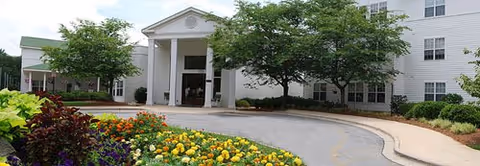 Front exterior view of The Carillon facility showing a white building with a covered entrance supported by columns, surrounded by trees and colorful flower beds along a curved driveway.