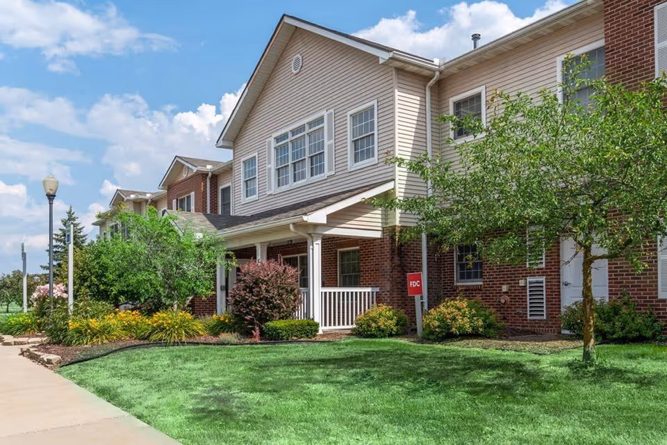 Front exterior of a two-story brick-and-siding senior living building with a covered porch, landscaped lawn, and sidewalk.