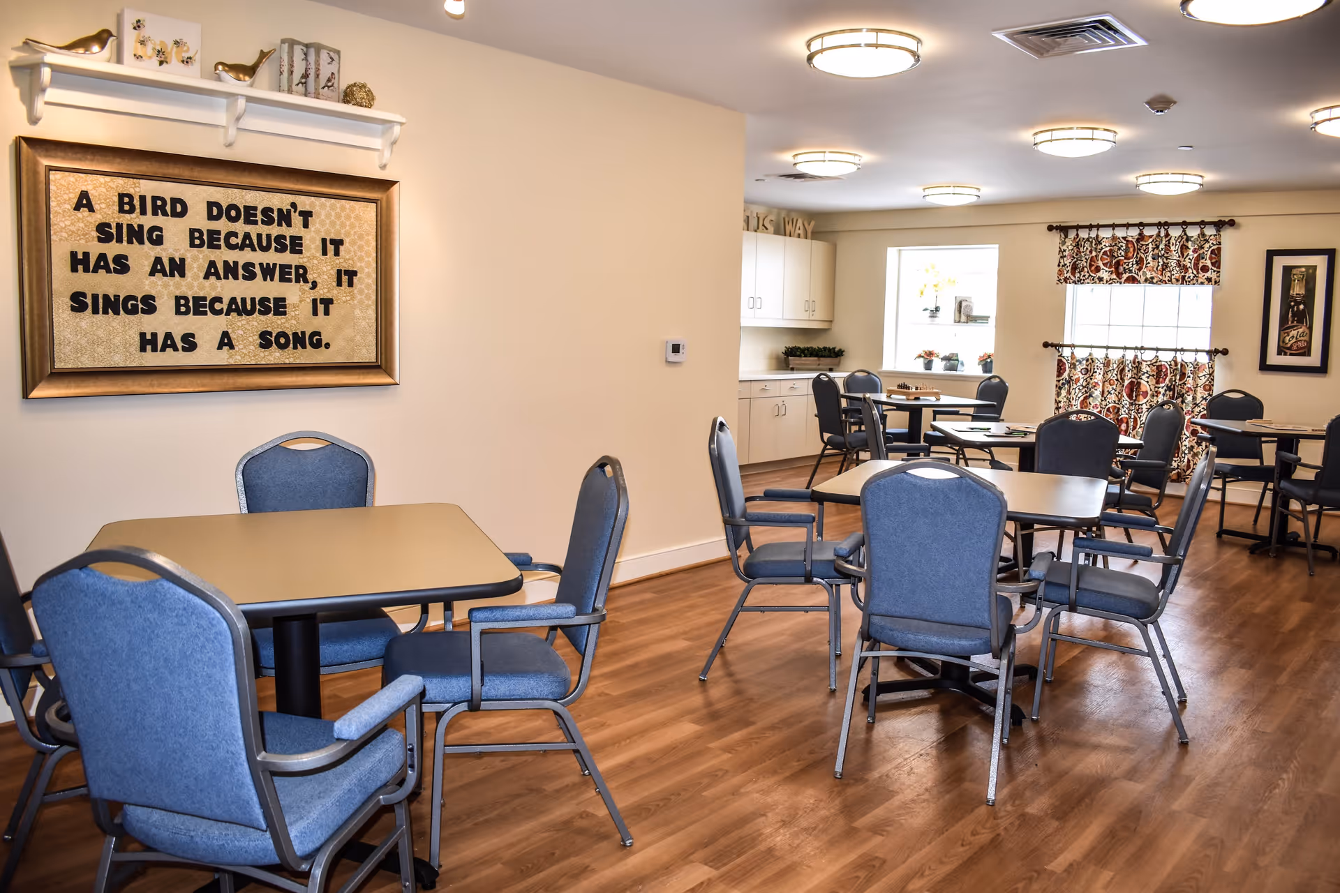 A well-lit dining room with several tables and blue cushioned chairs arranged around them. The room has wooden flooring, light-colored walls, and ceiling lights. On one wall, there is a framed quote that reads, 'A bird doesn't sing because it has an answer, it sings because it has a song.' There are windows with patterned curtains and a small counter with cabinets in the background.