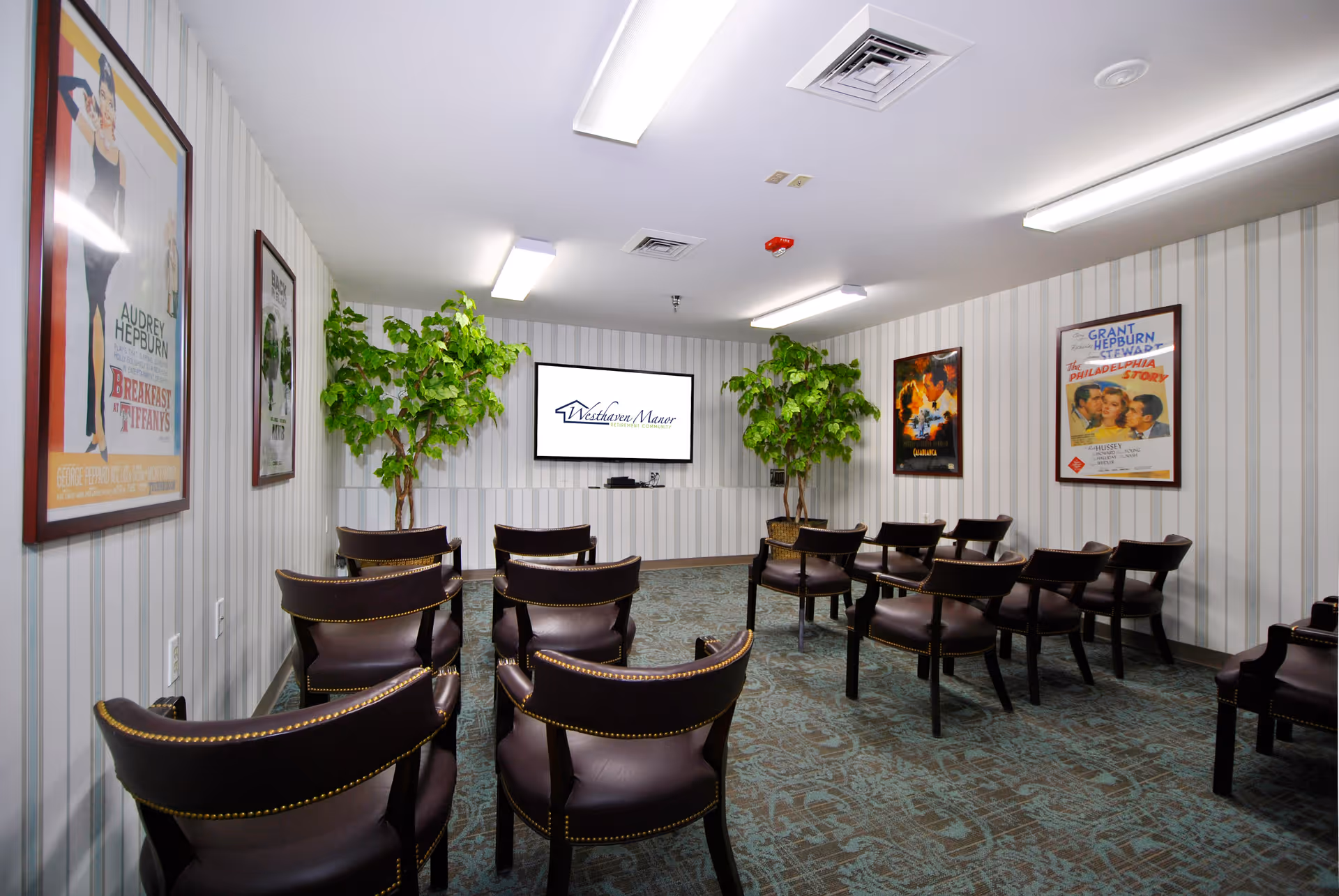 A small theater room with rows of brown leather chairs facing a wall-mounted TV screen displaying the Westhaven Manor logo. The room has striped wallpaper, framed classic movie posters on the walls, two large potted plants, and ceiling fluorescent lights.