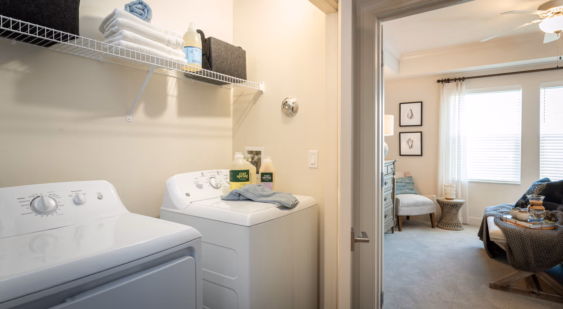 Laundry room with a white washing machine and dryer, a wire shelf above holding folded towels, cleaning supplies, and storage bins. The room opens into a cozy living area with a chair, side table, dresser, and large windows with white curtains.