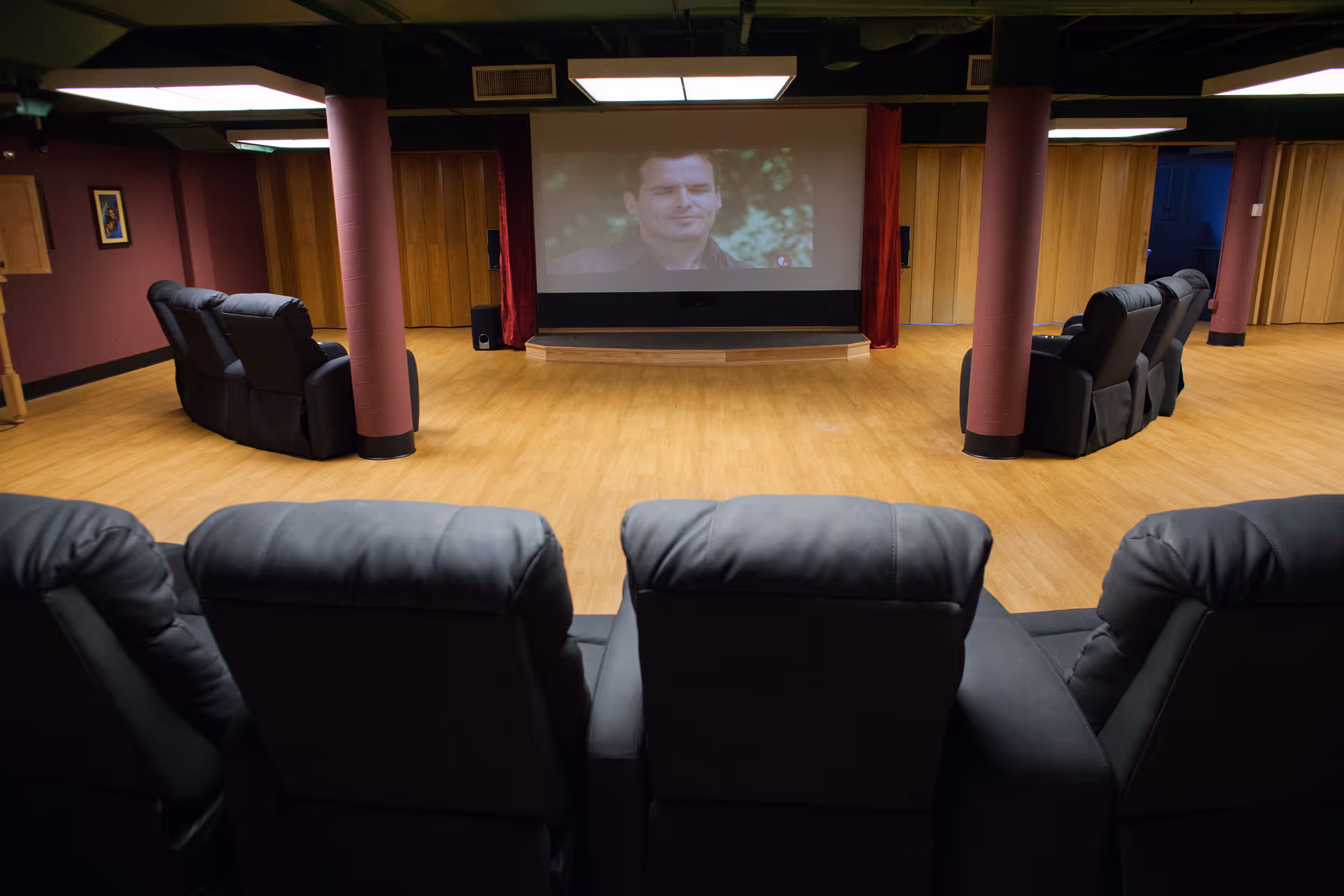 A cozy movie screening room with several black leather recliners arranged in rows facing a large screen showing a movie scene. The room has wooden flooring, maroon walls, and wooden paneling with two maroon columns near the screen.