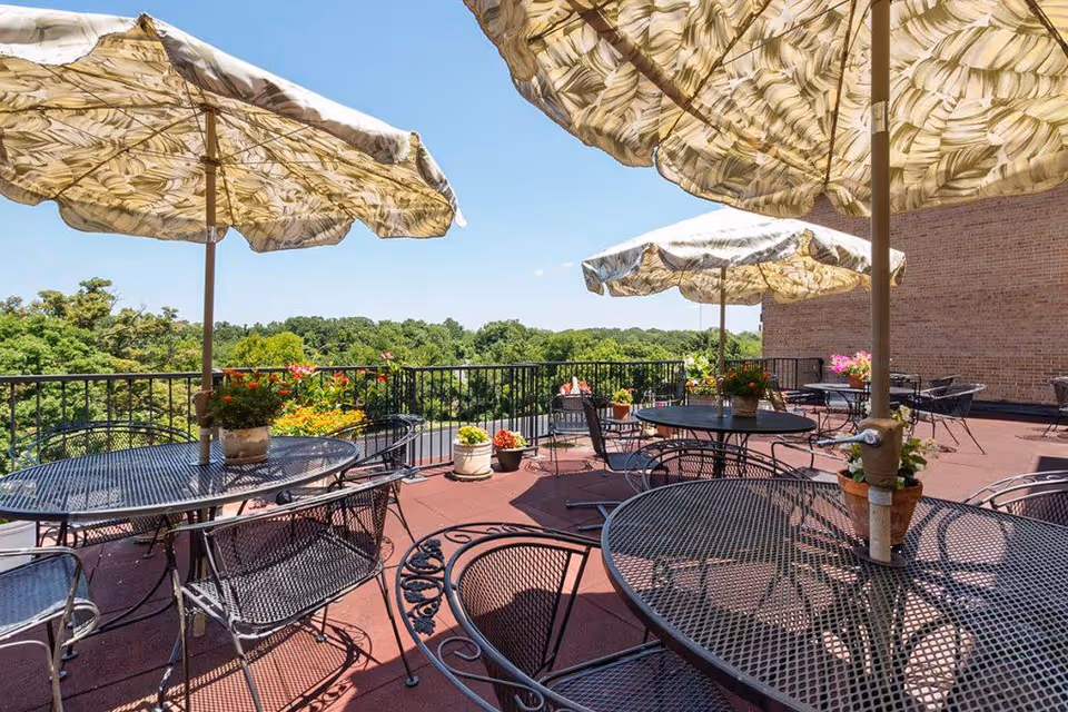 Outdoor patio area with several round metal tables and chairs, each table shaded by large patterned umbrellas. Potted plants and flowers decorate the space, with a view of green trees and a clear blue sky in the background.