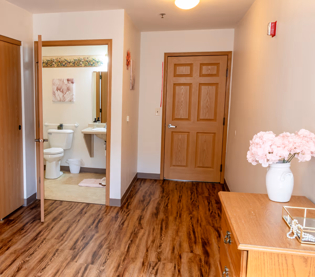 View of a hallway in a senior living facility with wood flooring and light-colored walls. There is a wooden door at the end of the hallway, and an open door on the left reveals a bathroom with a toilet, sink, and floral wall art. On the right side of the hallway, there is a wooden dresser with a white vase holding pink flowers and a decorative box with a pearl necklace.