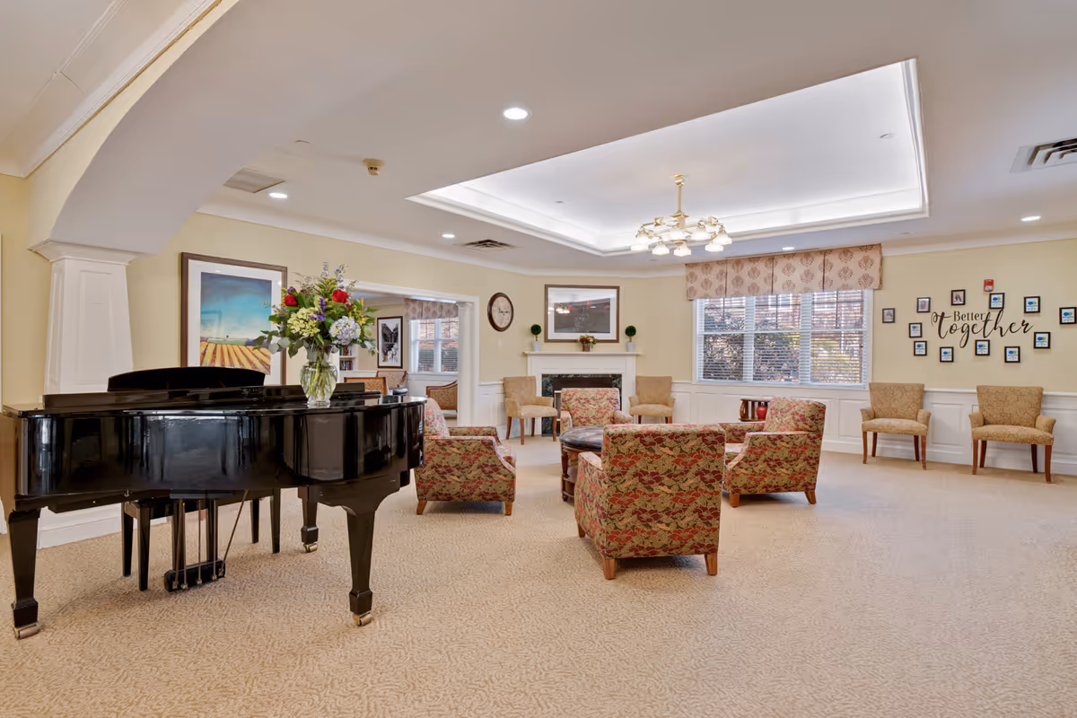 Spacious senior living common room with a black grand piano, floral upholstered chairs arranged around a coffee table, a fireplace, and large windows.
