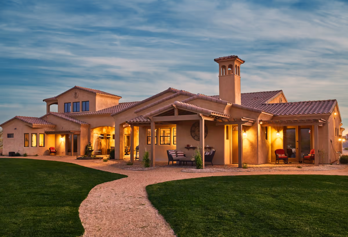 Exterior view of a large, single-story building with a southwestern architectural style, featuring stucco walls, a tiled roof, and outdoor seating areas. The building is surrounded by a well-maintained lawn and a gravel pathway leading to the entrance, under a partly cloudy sky at dusk.