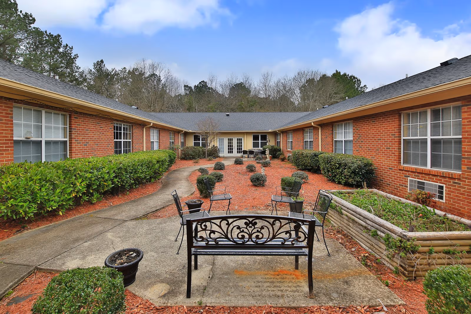 Outdoor courtyard area of a senior living facility with a paved walkway, metal chairs and benches, manicured bushes, and a raised garden bed. The courtyard is surrounded by a single-story brick building with multiple windows and a set of double doors at the far end. Trees and a partly cloudy sky are visible in the background.