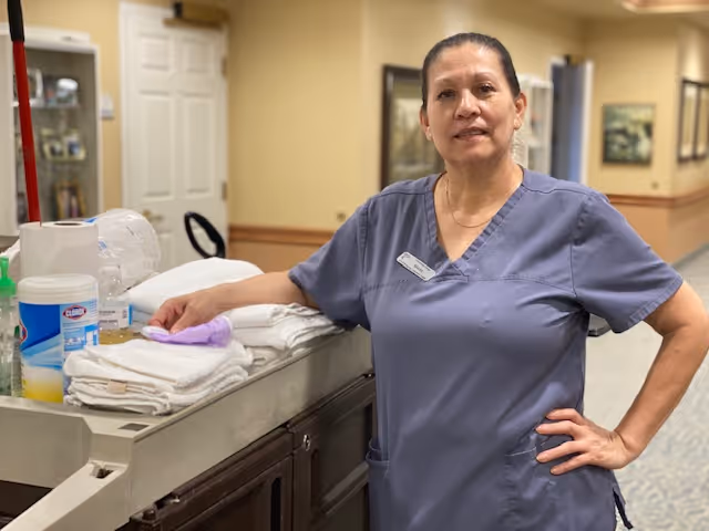 A woman in gray scrubs stands in a hallway of a senior living facility, leaning on a cart with folded towels and cleaning supplies. The hallway has beige walls with framed pictures and a carpeted floor.