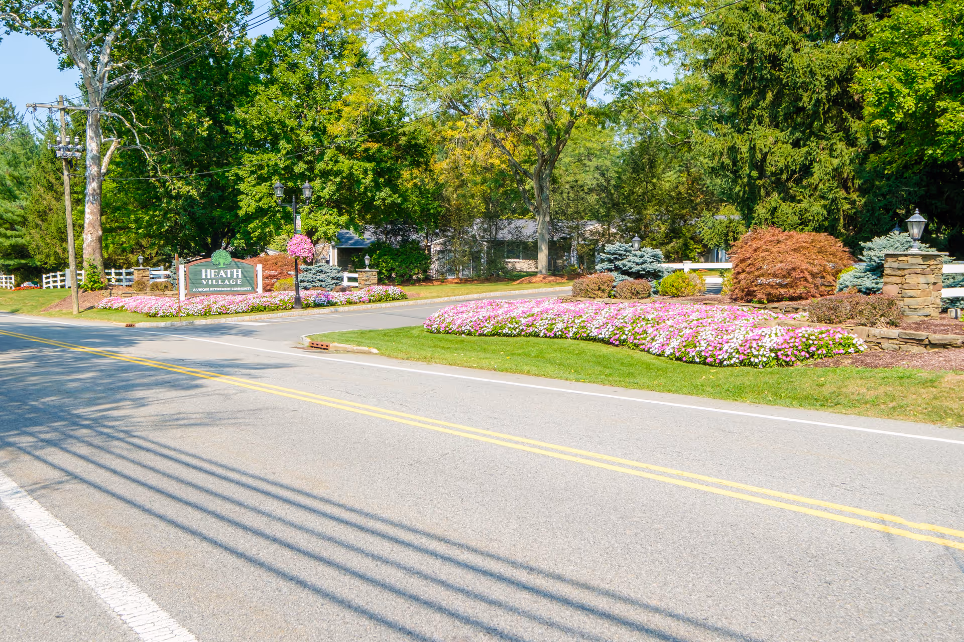 Entrance to Heath Village Retirement Community with a green sign surrounded by colorful flower beds, trees, and a paved road leading into the community.