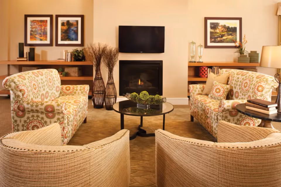 A cozy living room with patterned sofas, a round coffee table, and a fireplace. The walls are adorned with framed artwork, and decorative plants add a touch of greenery.