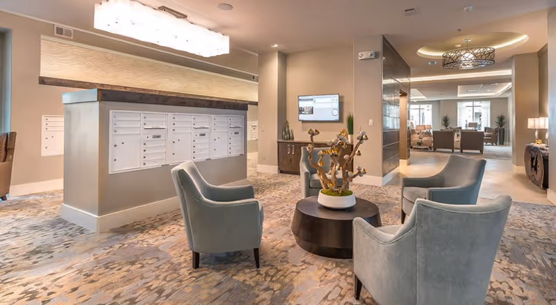 A modern and spacious interior common area with a cluster of four light blue armchairs arranged around a round dark coffee table holding a decorative plant. Behind the seating area is a large mailbox unit with multiple mail slots. The room features soft beige walls, patterned carpet flooring, recessed lighting, and a wall-mounted TV. In the background, additional seating areas and large windows letting in natural light are visible.