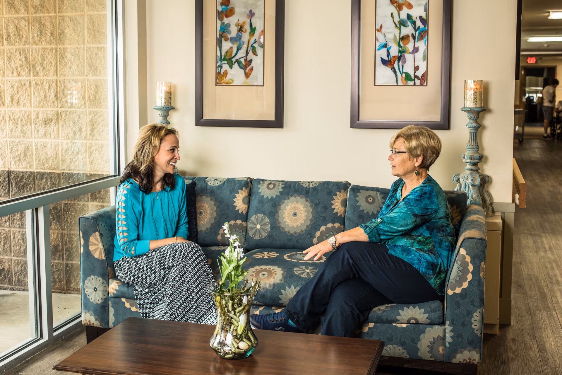 Two women sitting and talking on a patterned blue couch in a well-lit room with large windows. There are two framed floral artworks on the wall behind them and a wooden coffee table with a glass vase containing flowers in front of the couch.