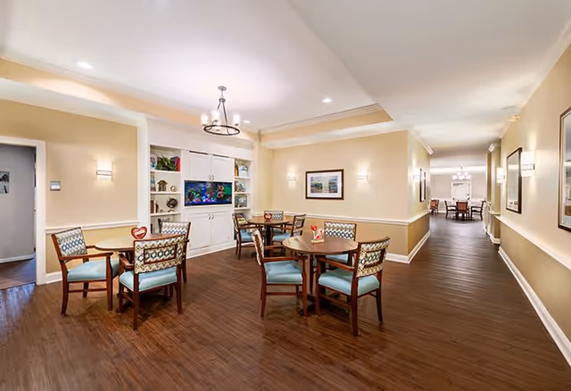 A well-lit senior living facility common area with several round wooden tables and cushioned chairs arranged for seating. The walls are painted in warm beige tones with white trim, and there is a built-in white cabinet with a television and decorative items. The floor is a dark wood laminate, and framed artwork hangs on the walls. The space extends into a hallway leading to another room with more seating visible in the distance.