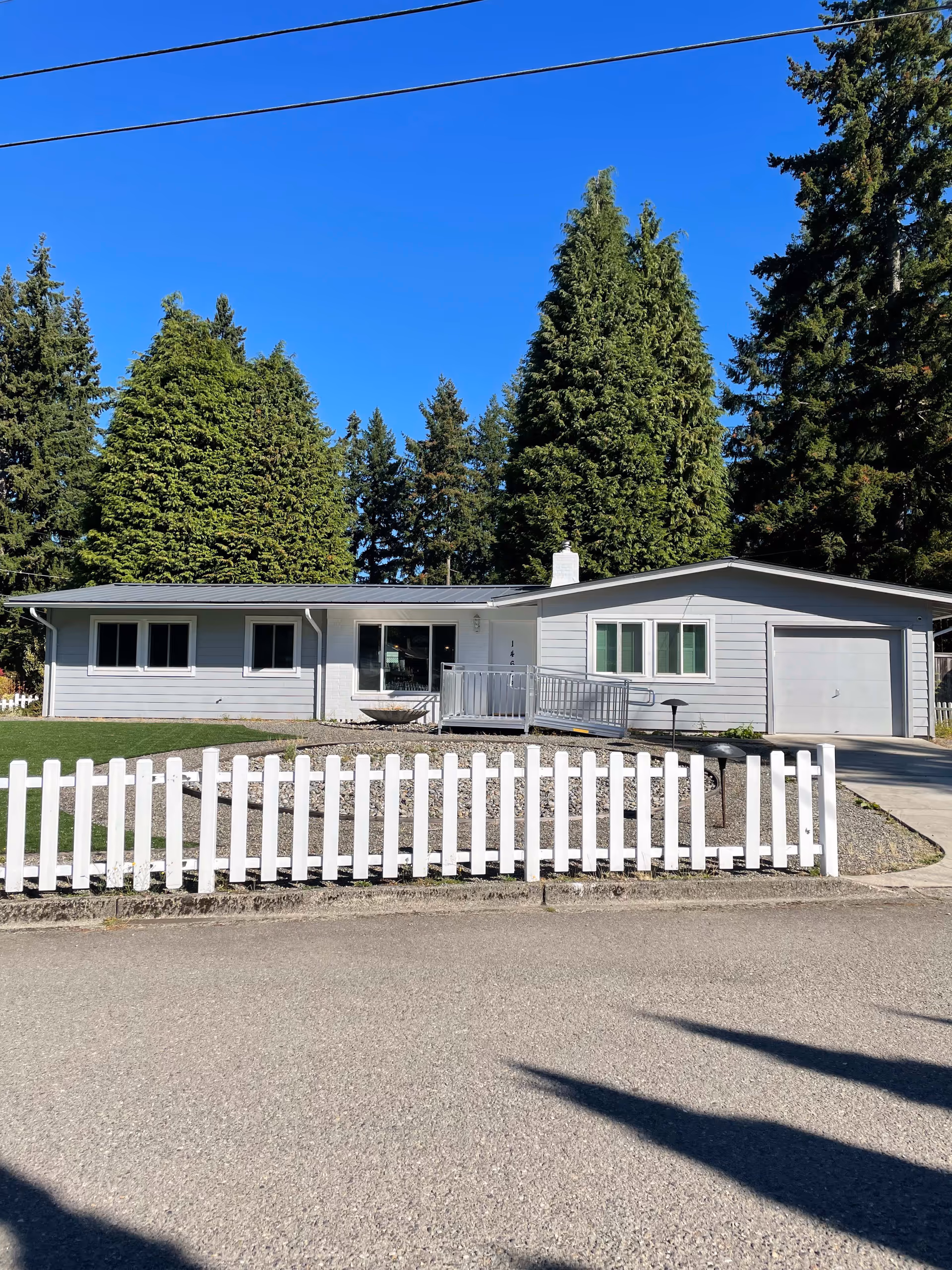Single-story house with light gray siding and a white picket fence in front. The house has several windows, a garage on the right side, a small ramp leading to the front door, and is surrounded by tall green trees under a clear blue sky.