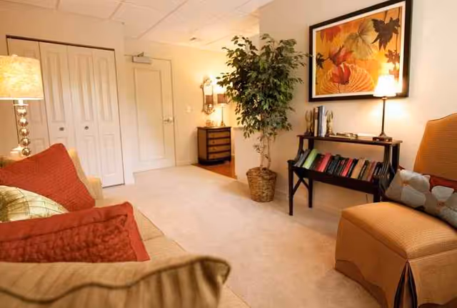 A cozy living room area with a beige carpet, a sofa with red and gold cushions, an orange armchair with a decorative pillow, a small black bookshelf filled with books, a large potted plant, a framed autumn-themed artwork on the wall, and warm lighting from a floor lamp and a table lamp.