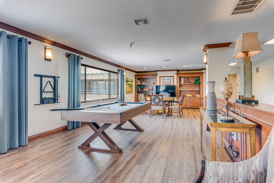 A spacious senior living common area featuring a pool table in the center, a table with chairs and a TV in the background, large windows with blue curtains, wooden flooring, and decorative lamps and vases on a console table along the right side.
