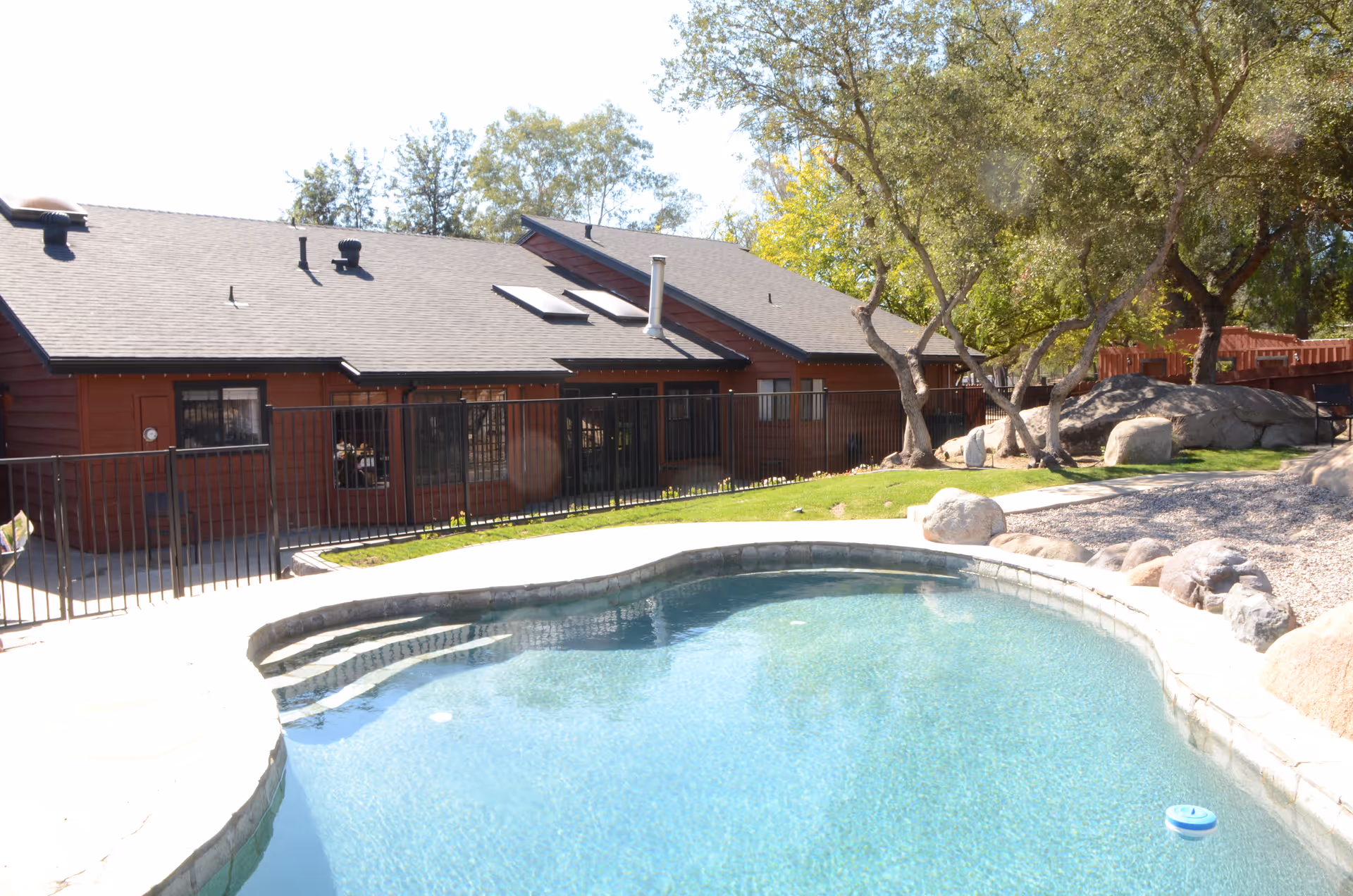 Outdoor view of a senior living facility showing a clear swimming pool in the foreground, a grassy area with rocks and trees, and a single-story building with a dark roof and red wooden exterior in the background.