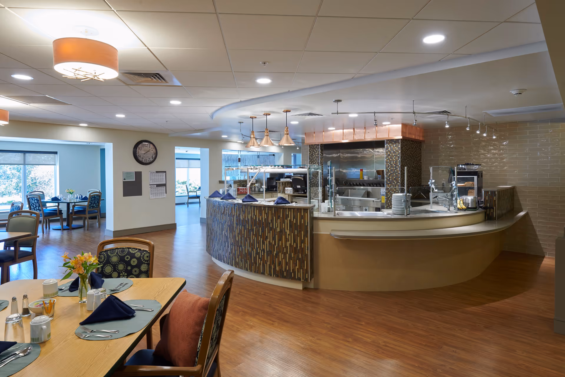 A dining area in a senior living facility with wooden floors, tables set with placemats, napkins, and utensils. The room features a curved serving counter with pendant lights above and kitchen equipment behind it. Large windows provide natural light to the space.