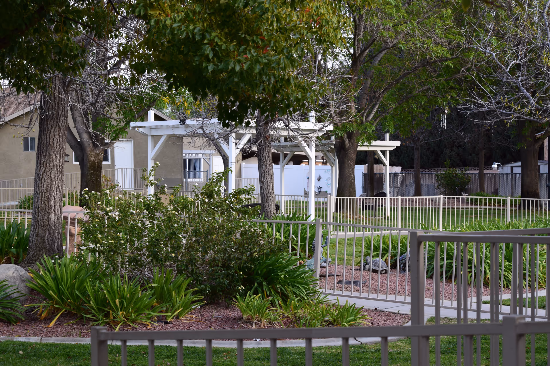 Outdoor garden area with green grass, trees, bushes, and a white pergola structure. There are white metal railings and a paved walkway surrounding the garden. In the background, there are buildings and a fence.
