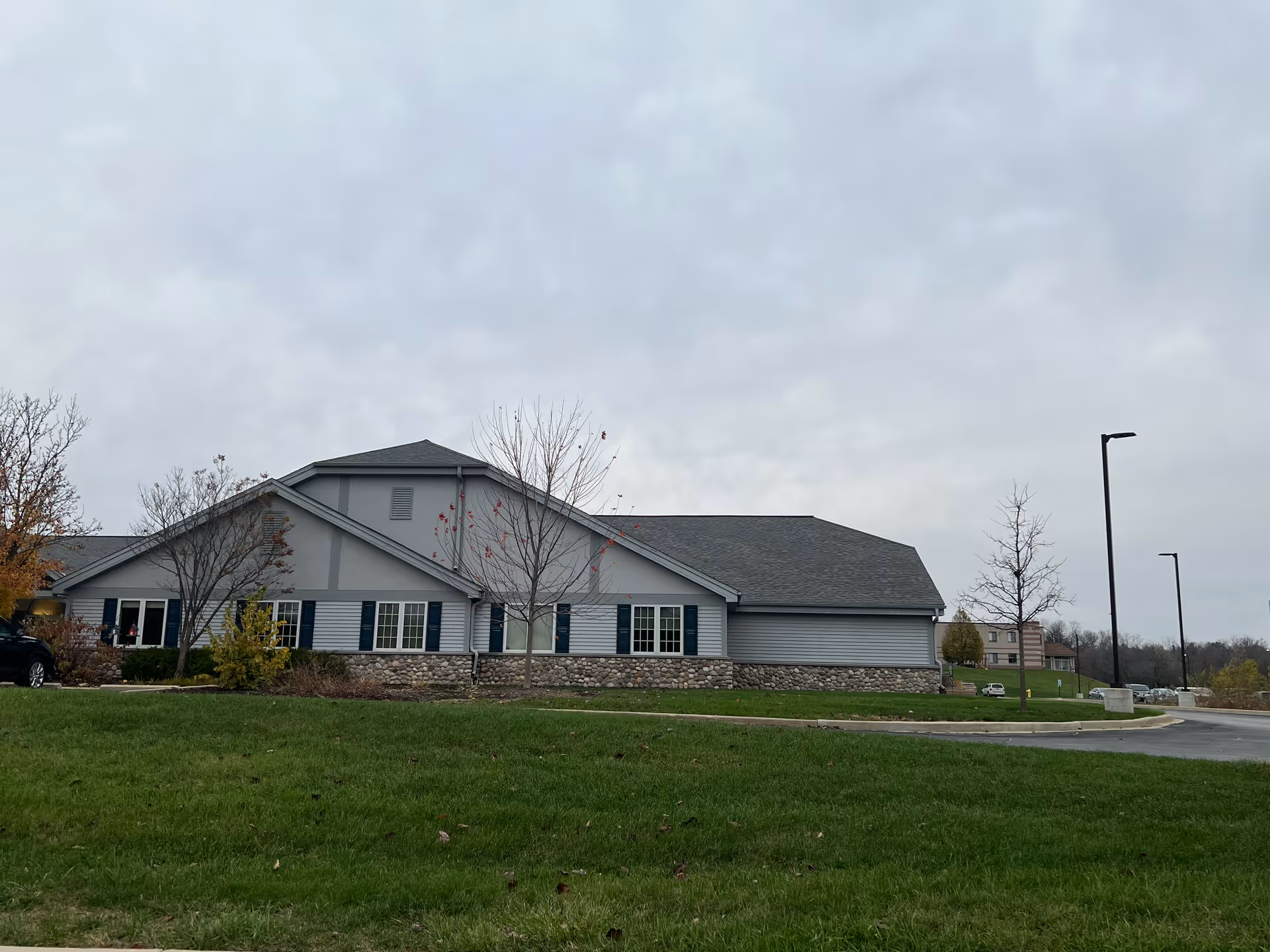 Exterior side view of a single-story building with gray siding, stone accents along the lower walls, and a dark gray roof. There are several windows with dark shutters and a few leafless trees in front of the building. The sky is overcast and there is a green lawn in the foreground with a paved driveway and street lamps to the right.