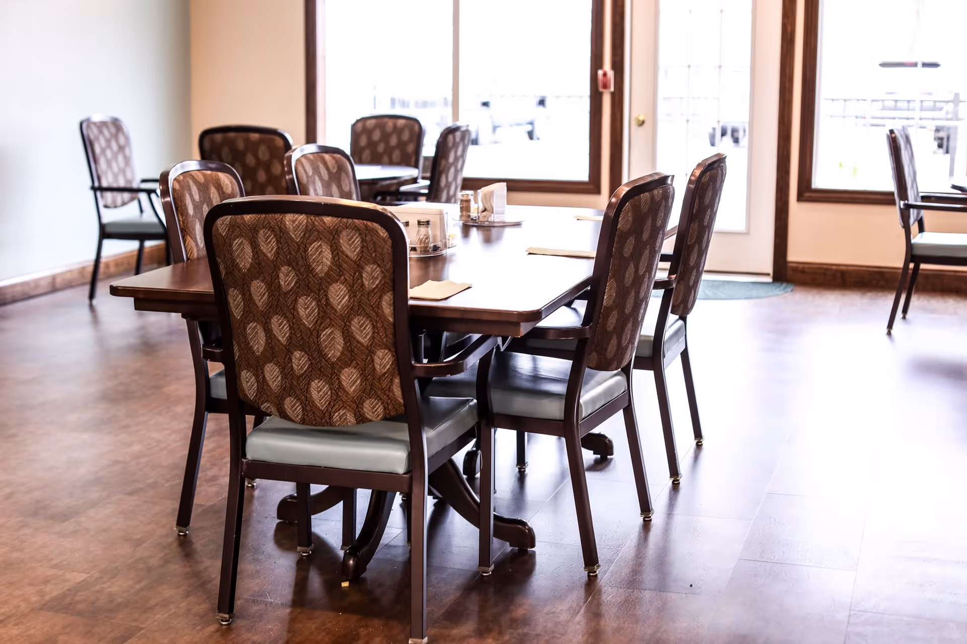 Dining area with wooden tables and upholstered chairs on a wood floor near large windows and a door.