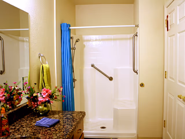 A bathroom with a granite countertop featuring a vase of pink and red flowers, a folded blue towel, and a bottle of soap. There is a large mirror above the countertop. The shower area has a white tiled wall, a built-in seat, a grab bar, and a blue shower curtain. A yellow towel hangs on a ring near the shower.