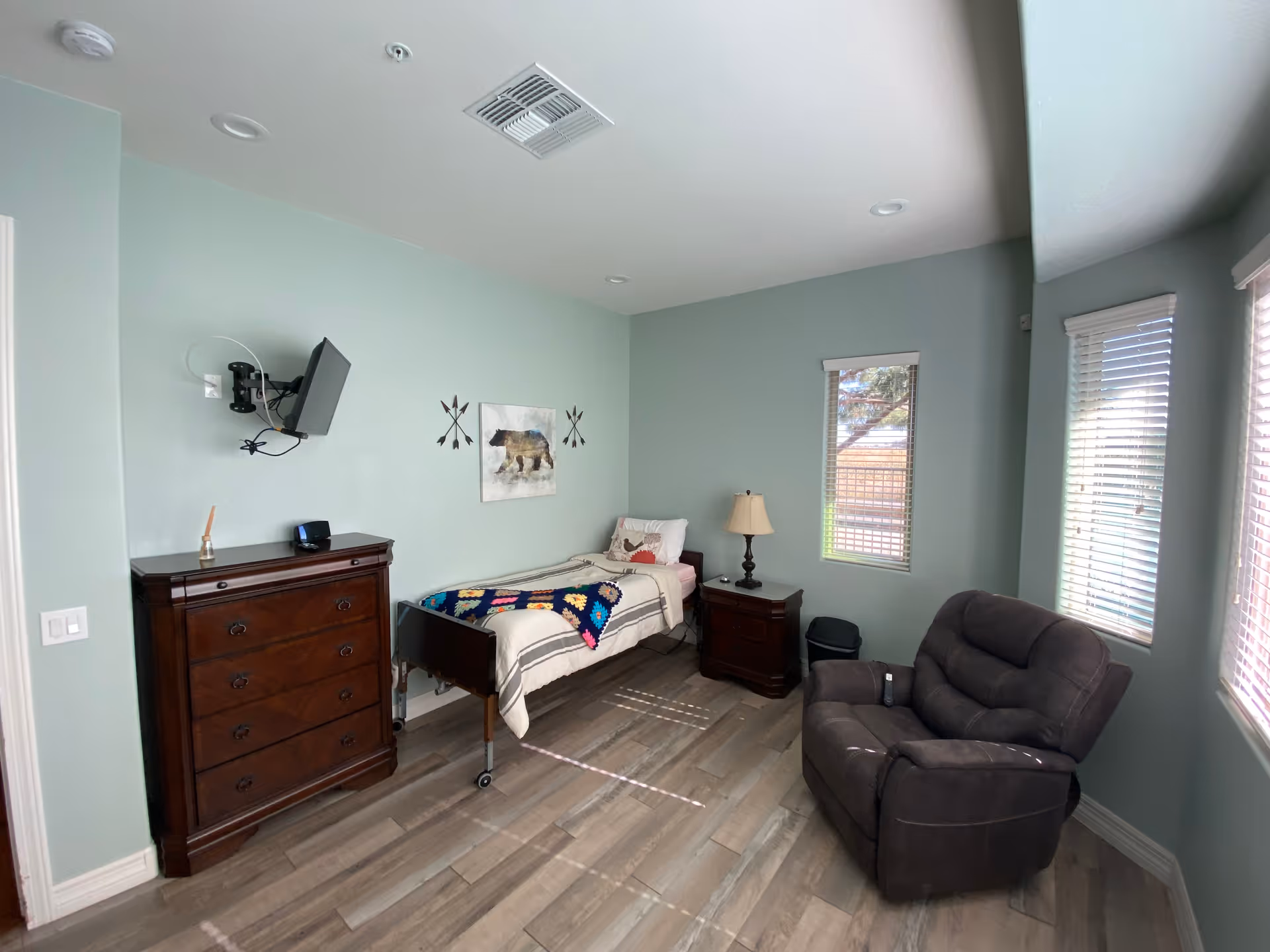 Bedroom in Eden Estates Assisted Living with bed, dresser, recliner, TV, and windows.